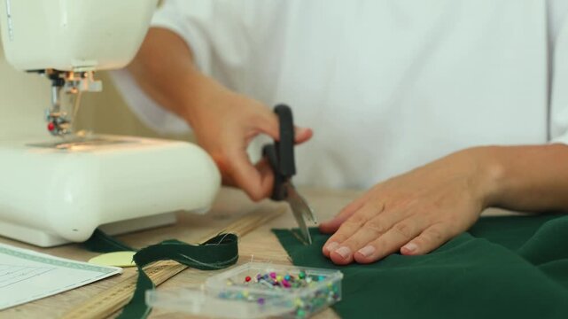 A person carefully cuts dark green fabric with scissors on a wooden table beside a sewing machine, pins, and pattern paper, preparing for garment construction.
