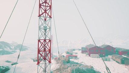 High above a snowy terrain, a communication tower stands tall near a cluster of red buildings. Snow blankets the ground, creating a serene but harsh atmosphere of isolation and research. © icetray