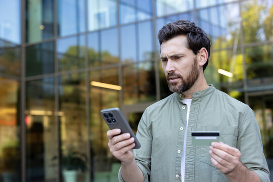 Man with worried expression holding smartphone and credit card outside a modern building, checking a declined payment or suspected fraud while online shopping and mobile banking