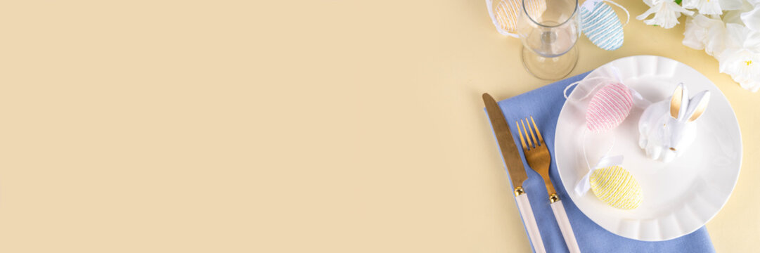 Cute Easter table setting with plate, cutlery set bunny, Easter egg, napkin and spring flowers on pastel colored yellow background, copy space, Easter brunch on dinner invitation, cafes menu layout