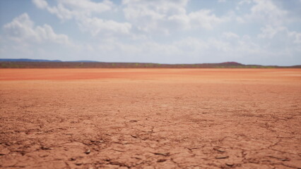 Fototapeta premium A vast, barren landscape stretches beneath a bright blue sky filled with soft clouds. The cracked earth reveals the harsh dryness of the land, with distant hills faintly visible.
