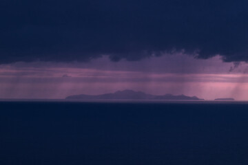 Fototapeta premium Cinematic wide shot of a distant island under a stormy purple sky. Features dramatic rain curtains and deep blue ocean. Ideal for travel, atmospheric, and background themes.
