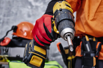 Construction worker wearing protective red work gloves and orange high-visibility overalls holds a yellow cordless electric drill. Professional power tools are strapped to his belt.