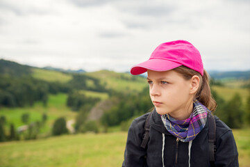 Thoughtful Teen Girl in Pink Cap, Plaid Scarf, and Backpack Looks Sideways over Lush Green Hills and Cloudy Sky