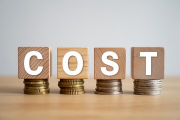 Four wooden blocks spelling out COST are stacked on coins representing business expenses