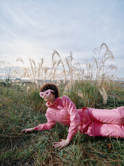 Obraz premium A person in a pink outfit lies on grass in an open field, relaxed pose among tall weeds, blue sky and soft light creating a casual outdoor portrait with nature