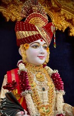 Obraz premium Beautiful close-up view of indian lord swaminarayan face glow, smile and blessing idol in the temple. isolated blurry red color designed costume, black curtains and gold interior view in background.