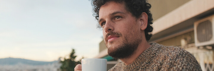Obraz premium Peaceful guy looks at the city from the balcony and drinks coffee, enjoys the beautiful view. Panoramic. Panoramic
