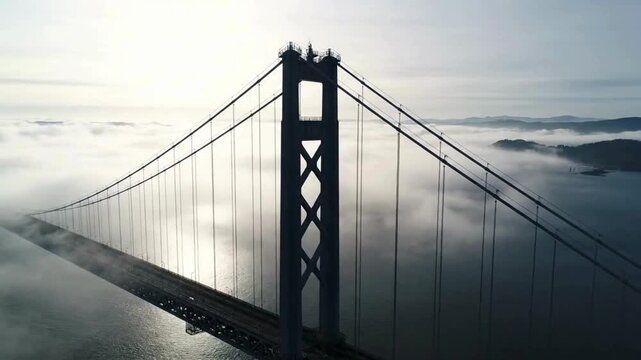 An aerial perspective of a majestic suspension bridge. The bridge gracefully spans across the water, enveloped in a serene embrace of mist and cloud, exuding an air of grandeur.