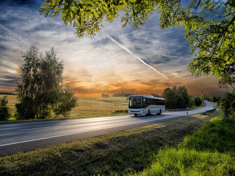 A white bus driving on the asphalt road in rural landscape at sunset