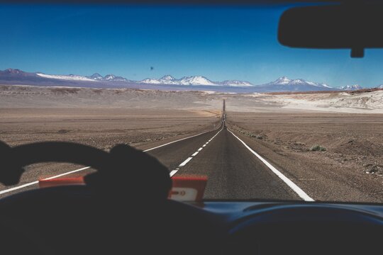 View from inside a vehicle driving along Ruta 23 across the Atacama Desert toward San Pedro de Atacama in northern Chile. The straight road cuts through the barren landscape.