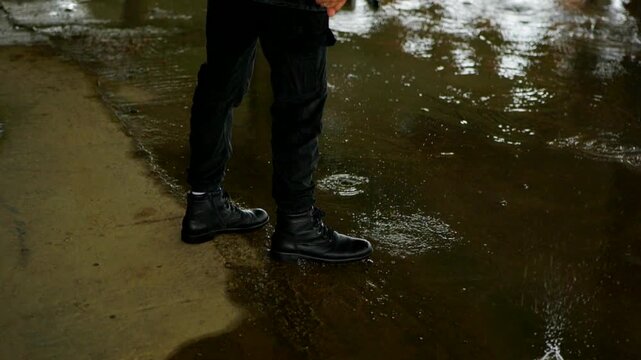 Black boots stand at the edge of a murky puddle, reflecting overhead light in the damp, urban setting. Close-up on dark trousers and lace-up boots near rippling water where raindrops create small spla
