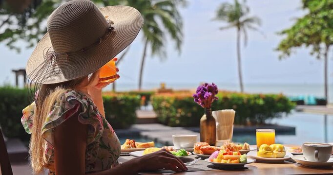 Woman wearing straw hat holding orange juice glass at poolside breakfast table, tropical ocean view with palm trees, bright sunny morning, luxury travel lifestyle, vacation relaxation.
