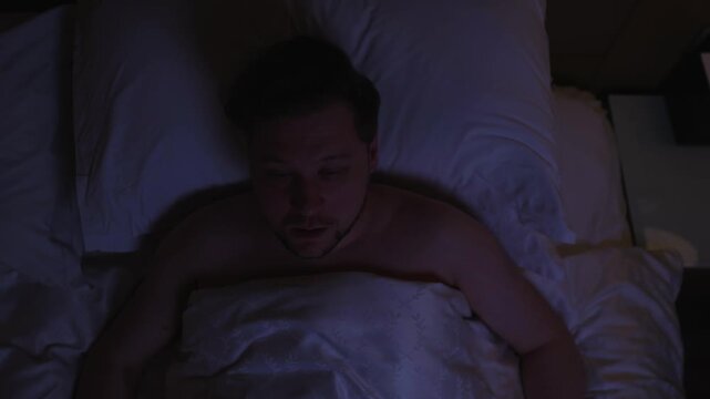 Frightened man suddenly waking up in his bed at night, clutching his chest and struggling to breathe, experiencing intense chest pain, panic, and fear during suspected heart attack in a dark bedroom