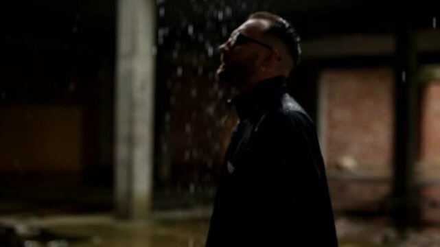 Man in profile, backlit by falling rain against a dark, moody backdrop. Silhouette of a man with slicked-back hair and glasses standing in the downpour. Dark, atmospheric shot of a man outdoors in the
