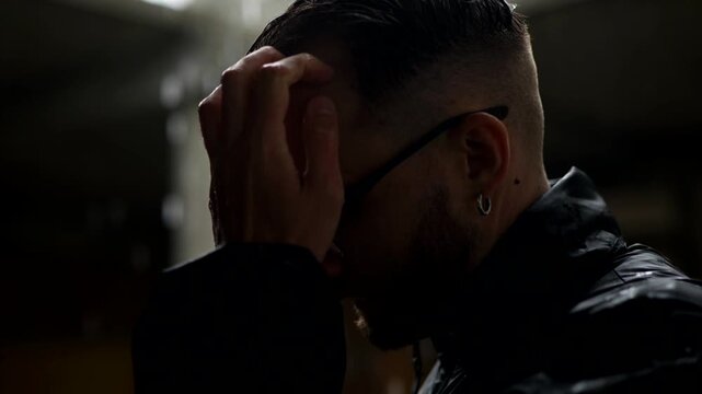 Man in profile, backlit by falling rain against a dark, moody backdrop. Silhouette of a man with slicked-back hair and glasses standing in the downpour. Dark, atmospheric shot of a man outdoors in the