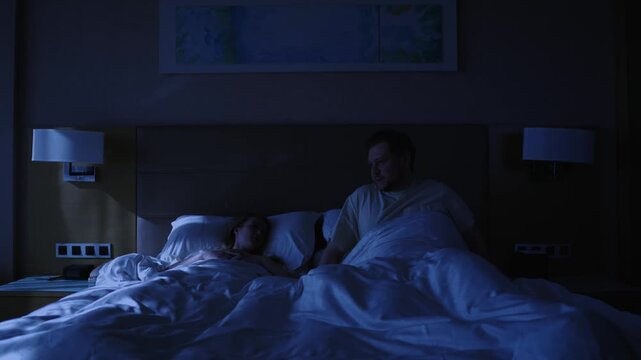 Mature man carefully getting into bed with his wife, who is already asleep, trying not to wake her up in dark bedroom late at night, finally settling down to rest after a long day