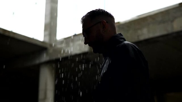 Man in profile, backlit by falling rain against a dark, moody backdrop. Silhouette of a man with slicked-back hair and glasses standing in the downpour. Dark, atmospheric shot of a man outdoors in the
