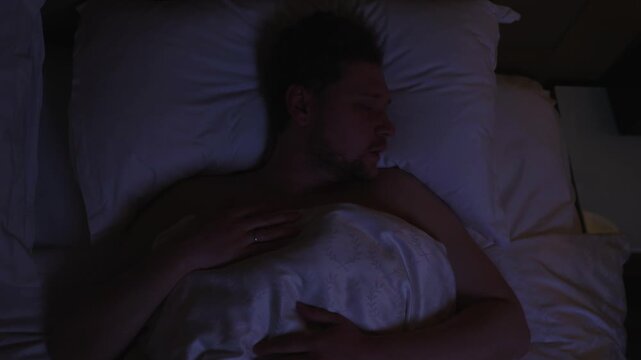 Top down shot restless man tossing and turning in bed at night, unable to sleep due stress, anxiety, or insomnia, rubbing his face and eyes in frustration, unable to sleep, struggling with insomnia
