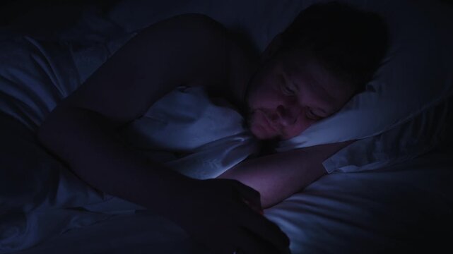 Sleepless man scrolling on his mobile phone in bed at night, with the screen light illuminating his face in the dark bedroom, showcasing issues with addiction and insomnia