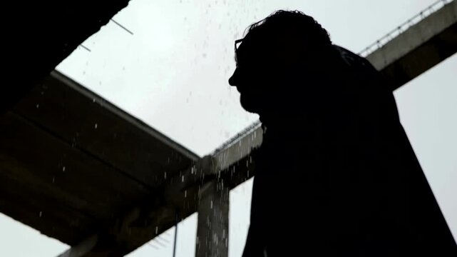 Man in black attire standing in a decaying, damp interior as water streams from the ceiling. Dripping water illuminates a dark, unfinished concrete space where a suited figure stands. Depressed man in