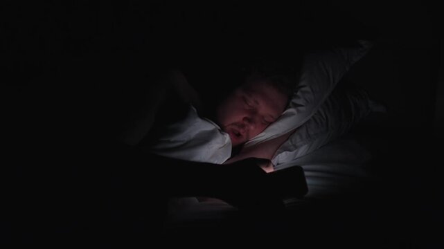 Sleepless man with insomnia doomscrolling on a mobile phone in a dark bedroom at night, the screen light illuminating his tired face as he lies on a pillow, unable to sleep in dark bedroom