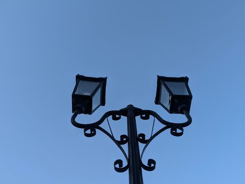 Low-angle view of a black, vintage-style double street lamp silhouetted against a bright, cloudless blue sky. The ornate metalwork and symmetrical design create a classic and elegant urban aesthetic. 