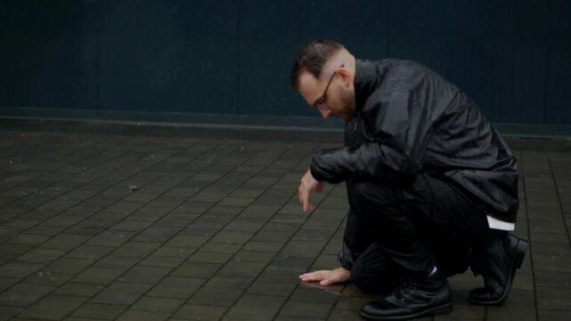 A man is kneeling under the pouring rain in the middle of the street against a black wall. A grieving man in despair under a heavy downpour. A powerless and desperate man stands on the asphalt.