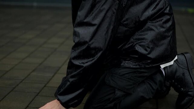 A man is kneeling under the pouring rain in the middle of the street against a black wall. A grieving man in despair under a heavy downpour. A powerless and desperate man stands on the asphalt.