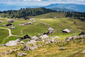 Naklejka premium A wide panoramic view of a traditional shepherd village with unique wooden huts scattered across green rolling hills under a dramatic sky in Velika Planina, Slovenia.