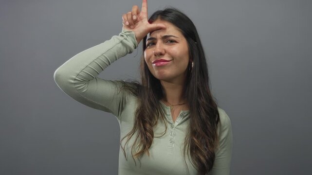 Young hispanic woman making loser gesture with hand on forehead against a grey wall in studio session; mockery.