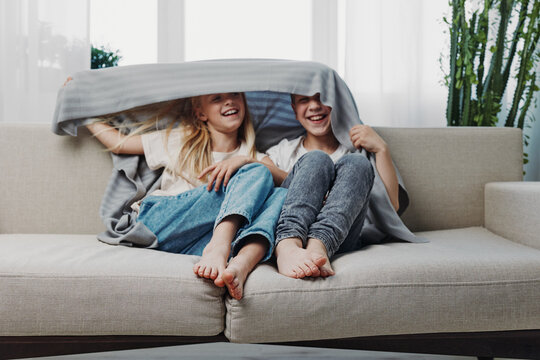 Two happy children sitting barefoot on sofa smiling and playing hide and seek under blanket in bright living room. Concept of family fun, childhood joy, and cozy home moments.