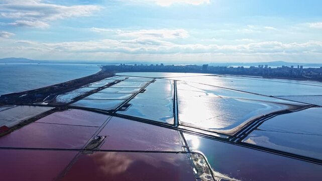 A stunning view of expansive salt flats shimmering in shades of pink and blue, capturing sunlight as it dances across the water with a coastal city rising in the distance.