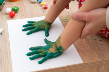 Child creates handprint art during a craft session in a home setting, surrounded by paint and materials on a wooden table