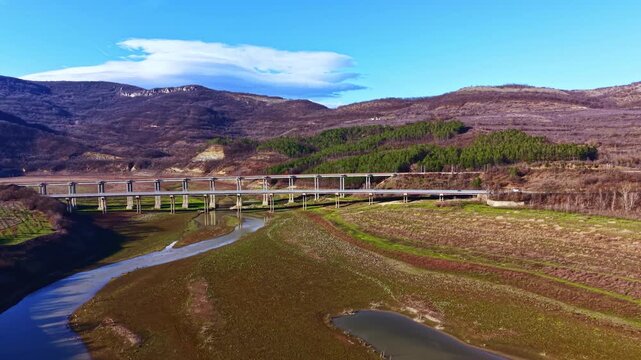 A bridge spans across a river in a valley. Hills rise in the background with patches of green trees. The landscape shows dry ground and few plants, while the sky is blue with some clouds.