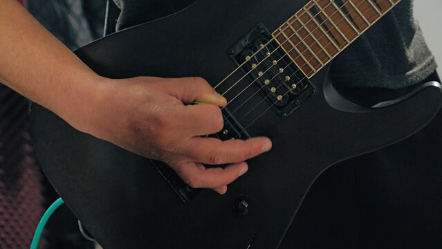 A close-up action shot of a rock musician's hand strumming a matte black electric guitar. The focused, dynamic composition captures the energy and passion of a rehearsal or live performance