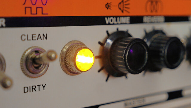Close-up of a retro guitar amplifier. A warm, glowing light shines next to the clean dirty channel switch, with volume knobs blurred in the background. Ideal for music and audio concepts