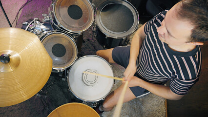 Obraz premium High-angle shot of a young man playing the drums with passion. He is focused on his performance during a band rehearsal in a music studio with purple stage lights creating an energetic atmosphere