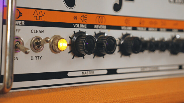 Detailed close-up of a retro guitar amplifier. The shot has a shallow depth of field focusing on the knobs and switches. A warm orange light glows, indicating the amp is on. Ideal for music themes