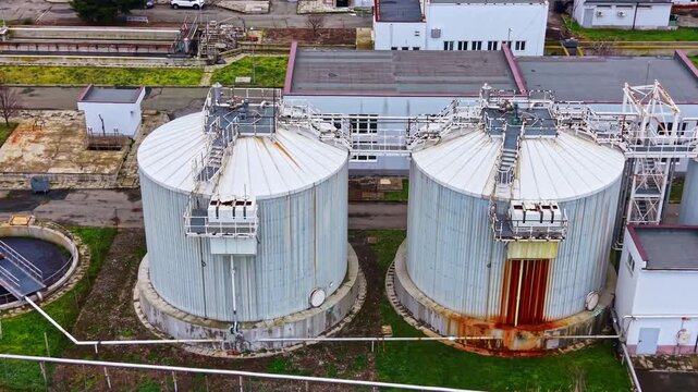 Two large storage tanks stand at an industrial location with surrounding buildings and a clear sky above. The setting shows signs of use and age.