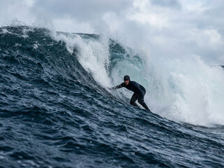 A lone surfer in a black wetsuit riding a large, powerful ocean wave under an overcast sky.