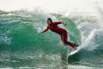A female surfer in a red wetsuit rides a powerful green wave, expertly maneuvering her board with spray flying around her.