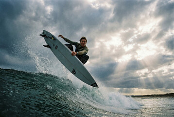 Surfer performing an impressive aerial maneuver on a wave under a dramatic cloudy sky with sun rays.