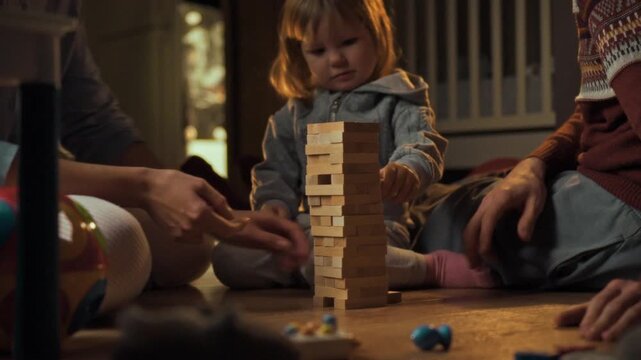 In warm cozy home setting, young family, including mom, dad, their little daughter, gather on living room floor for fun-filled Jenga game, bonding and laughing together amidst comfortable surroundings