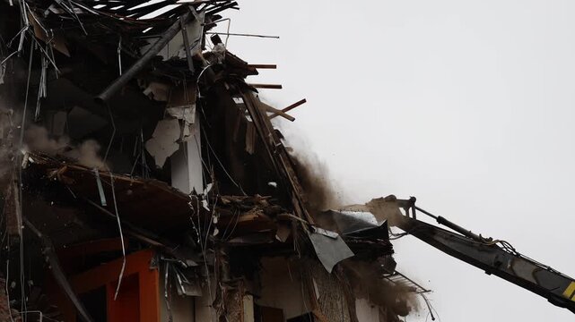 Excavator arm with hydraulic shears tears into a partially collapsed building, ripping concrete and steel as dust and debris billow, exposing beams and interior panels during an intense teardown