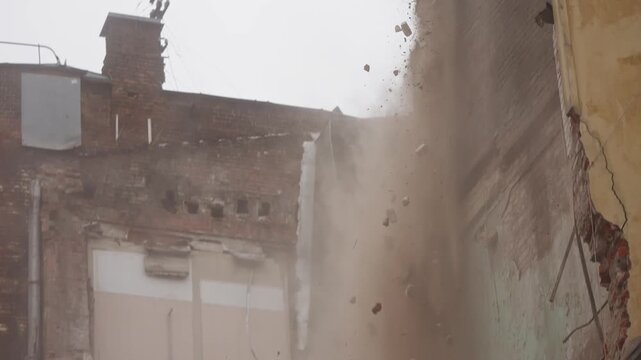 Excavator hydraulic arm demolishes a crumbling brick wall, sending clouds of dust and debris across an industrial site as the structure collapses amid rubble and shattered masonry