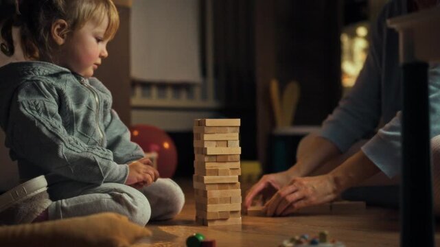 Young daughter her mother playfully construct precarious Jenga tower on floor during warm evening, fostering family bonding interactive learning through balanced entertainment and fun board game play
