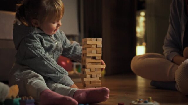 A young daughter and mother spend time together playing Jenga, building a wobbly tower. A warm evening in cozy setting encourages interactive entertainment and shared learning through playful laughter