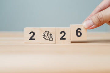 Wooden blocks arranged to show 2026 with hand adjusting the final number, featuring globe and thermometer icon symbolizing climate change, global warming and future environmental impact on background.