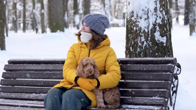 Young woman with brown hair in yellow winter coat sits on a bench holding a small dog, surrounded by snow-covered trees in a winter park setting
